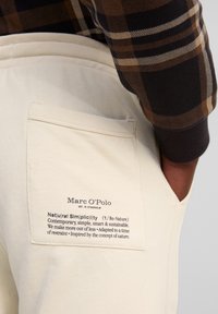 Beige Baumwollhosen mit einer Gesäßtasche, die ein bedrucktes Etikett aufweist. Das Etikett enthält den Markennamen und Texte zur Nachhaltigkeit.