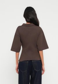 Chemise marron à manches courtes avec un col plat, une coupe légèrement cintrée et une texture douce, portée avec un jean foncé.