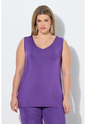 Frau mit blondem Haar, die ein ärmelloses lila Oberteil und passende Hosen trägt, dazu lange gold-schwarze Ohrringe, auf weißem Hintergrund.