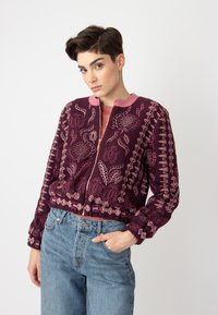 Veste bomber bordeaux brodée avec des accents roses, ornée de motifs floraux et dotée d'une fermeture éclair, associée à un jean bleu clair taille haute.
