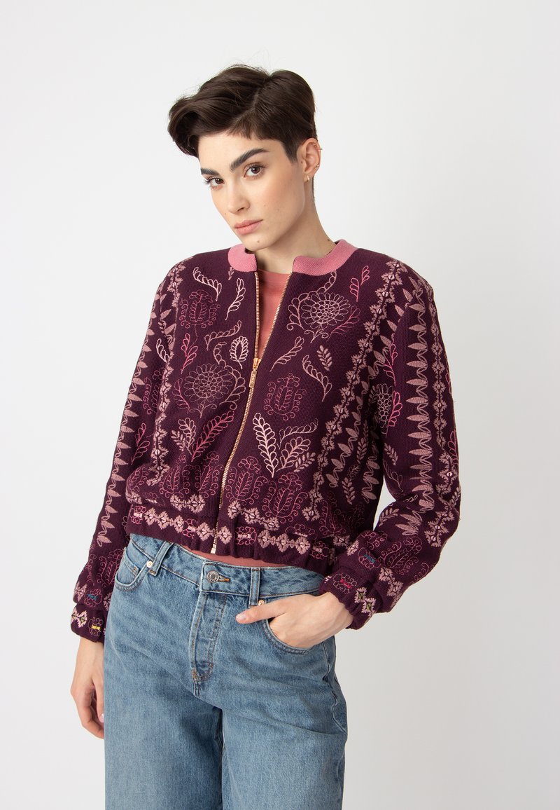 Veste bomber bordeaux brodée avec des accents roses, ornée de motifs floraux et dotée d'une fermeture éclair, associée à un jean bleu clair taille haute.