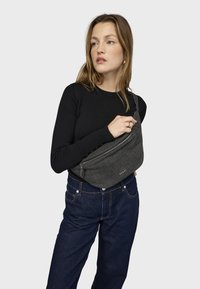 Sivý ľadvinkový pás s vreckom na zips a plochým dizajnom, nosený cez telo na čiernom tričku s dlhým rukávom a modrých džínsoch.