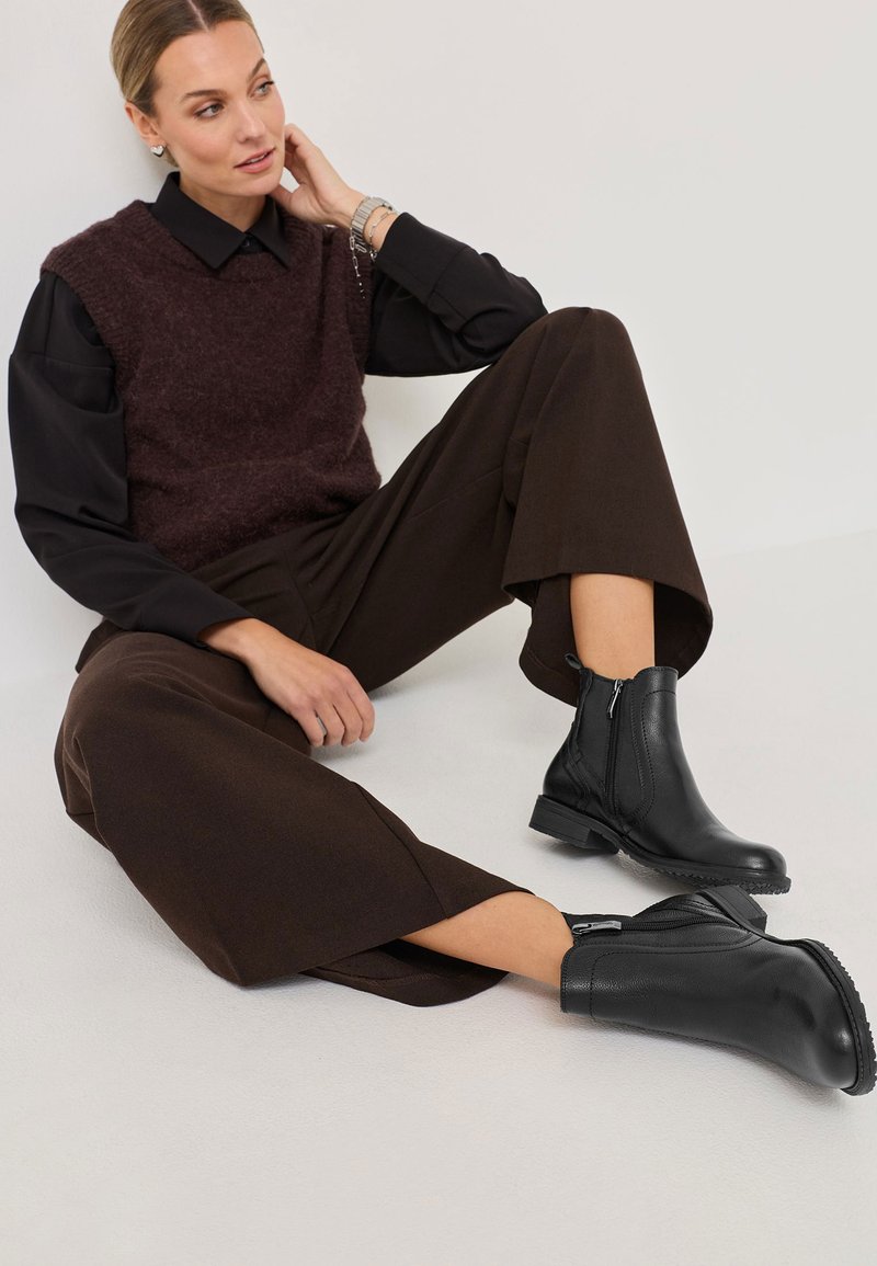 Černé kožené kotníkové boty se zipem po straně, s texturovaným povrchem. Nosené s tmavě hnědými kalhotami širokého střihu a burgunďským svetr vestou.