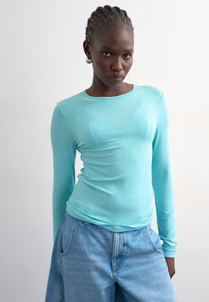 Osoba s copánky, která nosí stříbrné kruhové náušnice, tyrkysovou košili s dlouhými rukávy a volné světle modré džíny na bílém pozadí.