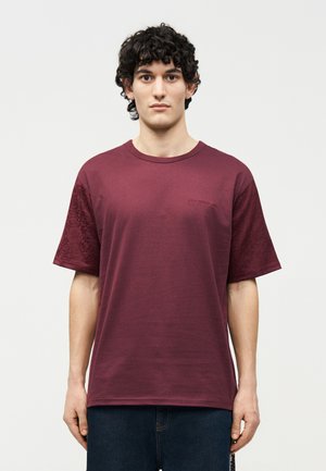 Fiatal férfi göndör sötét hajjal, sima bordó rövid ujjú pólót és sötét farmert viselve, fehér háttér előtt áll.