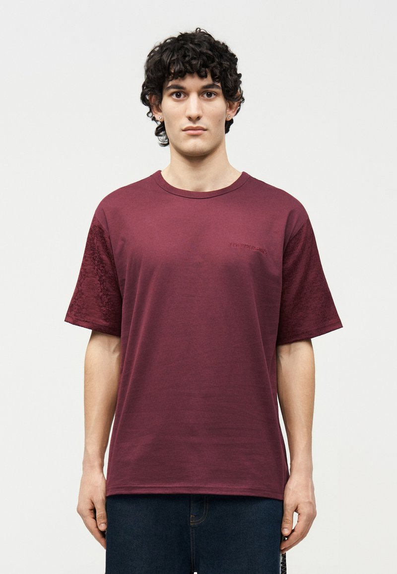 Νέος άνδρας με σγουρά σκούρα μαλλιά, φορώντας μια απλή μαρουν κόκκινη κοντομάνικη μπλούζα και σκούρα τζιν, στέκεται μπροστά σε ένα λευκό φόντο.