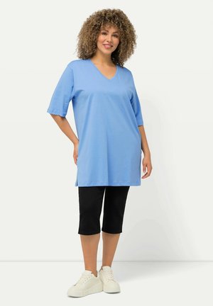 Helle blaue V-Ausschnitt-Tunika mit kurzen Ärmeln, kombiniert mit schwarzen kurzen Hosen und weißen Turnschuhen. Glatte Baumwolltextur, lässiges Design.