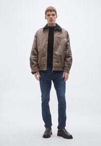 Καφέ μπουφάν τύπου bomber με μαύρο γιακά, τσέπη με φερμουάρ, φορεμένο πάνω από μαύρο πλεκτό πουλόβερ με ρίγες, συνδυασμένο με μπλε τζιν και μαύρα παπούτσια.