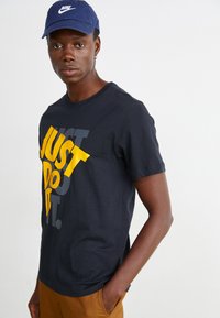 T-shirt de algodão preto com grande gráfico amarelo e cinza "JUST DO IT." Mangas curtas, gola redonda, textura suave. Combinado com calças castanhas.