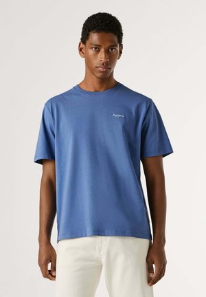 Homme portant un t-shirt bleu uni à manches courtes et un pantalon blanc, debout devant un fond clair, faisant face à l'avant avec une expression neutre.