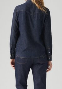 Mörk denimskjorta med lång ärm, knappmanschetter och en rundad nederkant. Den har sömnad på baksidan och ljusa kontrastsömmar.