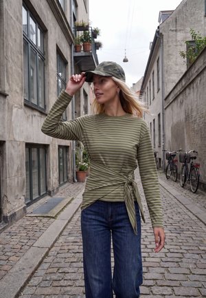 Une femme ajuste sa casquette de camouflage dans une rue pavée bordée de vieux bâtiments et de vélos stationnés.