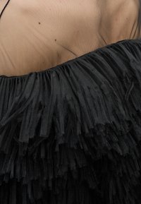 Κοντινό πλάνο ενός ατόμου που φοράει μαύρη φούστα με φράντζες και διάφανο μπεζ τοπ από δίχτυ.