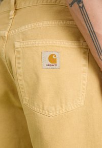 Geltonos džinsinės kelnės su užpakaline kišene, ant kurios yra kvadratinė Carhartt logotipo etiketė. Medžiaga turi lygią tekstūrą su subtiliu įstrižiniu audimu.