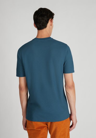 Hombre con cabello corto y oscuro, vistiendo una camiseta azul de manga corta sencilla y pantalones de pana naranjas, de pie de espaldas contra un fondo liso.