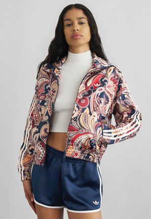 Kvinde iført en farverig jakke med paisley-mønster med hvide striber og marineblå Adidas-shorts med hvid kant, stående med den ene hånd på hoften.
