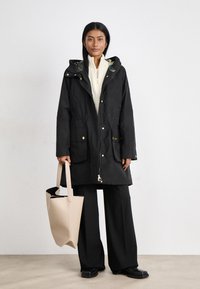 Zwarte waterdichte parka met een capuchon, twee voorkantenzakken, gouden accenten en een ritsluiting. Model houdt een beige tote bag vast.
