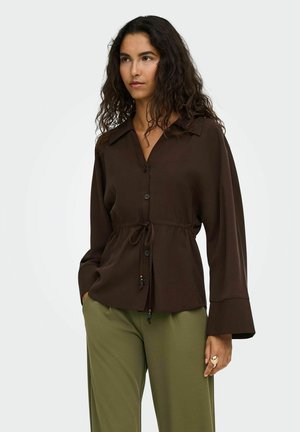 Femme aux longs cheveux bouclés portant une chemise boutonnée marron foncé avec une taille à cordon et un pantalon vert olive.