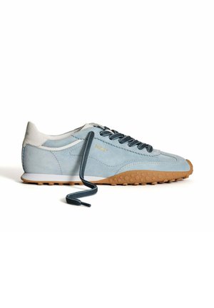 Zapatilla de ante azul claro con talón blanco y suela de goma marrón. Presenta cordones azul oscuro y acentos texturizados en la suela. Detalle del logo en dorado.