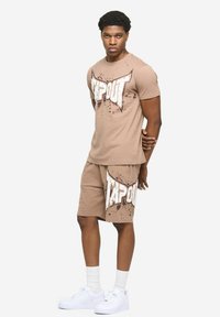 Bež majica kratkih rukava i odgovarajuće kratke hlače s upečatljivim logotipom "TAPOUT" s efektima prskanja. Bijele tenisice i čarape upotpunjuju odjevnu kombinaciju.