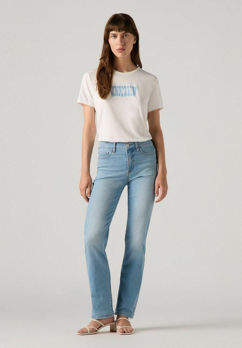 Jeans de color azul claro con corte recto, combinados con una camiseta blanca que presenta un logo "Levi's" en azul claro. El modelo lleva sandalias de dedo.