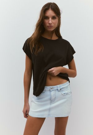 Giovane donna che indossa una maglietta nera larga e una minigonna di denim azzurro chiaro, solleva leggermente la maglietta per mostrare la pancia.