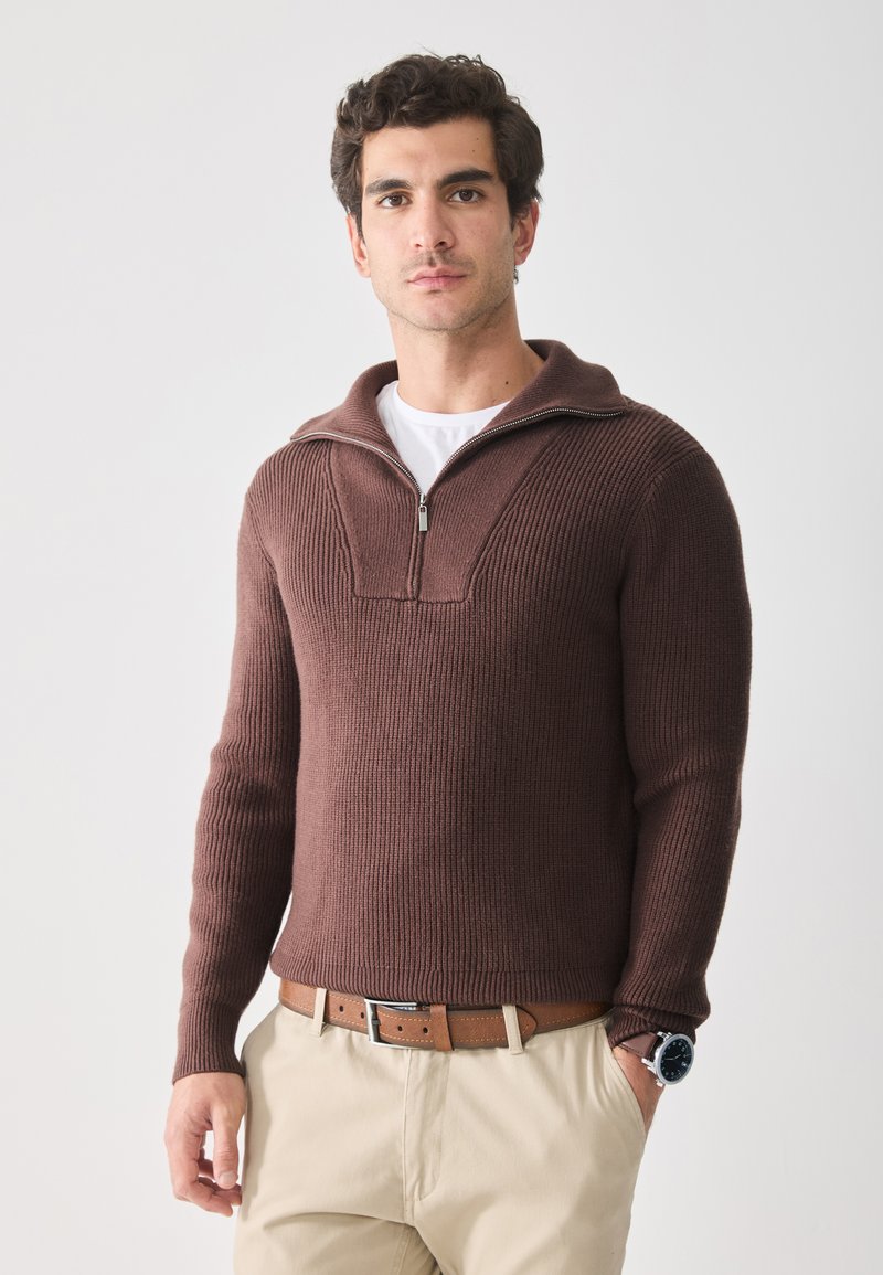 Pull en maille côtelée marron avec un col zippé, présentant un motif vertical et porté sur une chemise blanche, associé à un pantalon beige et une ceinture.