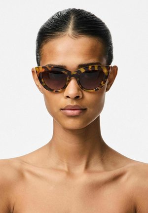 Femme aux cheveux lisses tirés en arrière, portant de grandes lunettes de soleil œil de chat écaille de tortue, expression neutre, épaules nues, fond blanc.