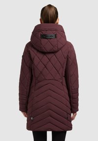 Chaqueta acolchada marrón con capucha, que presenta un patrón en forma de chevron en la parte posterior y un parche con el logo en el cuello. Textura suave y diseño cosido.