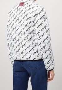 Vit puffjacka med ett upprepande svart "Hugo"-logomönster, med en vinröd etikett vid kragen och en strukturerad, quiltad design.