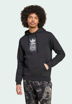 Sudadera negra con capucha que presenta un estampado gráfico en el frente y un bolsillo tipo canguro, combinada con pantalones oscuros de patrón. La tela parece suave y texturizada.