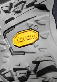Szara gumowa podeszwa z głębokimi bieżnikami i wyraźnym logo Vibram w kolorze żółtym. Teksturowana powierzchnia i kątowe wzory poprawiają przyczepność i trakcyjność.