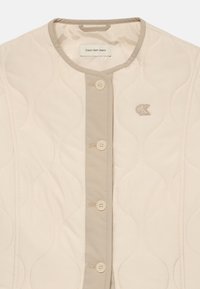 Beige quiltet jakke med rund hals, knaplukning, kontrastbesætning og lille broderet logo på venstre bryst.