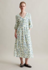 Das florale Midikleid hat einen V-Ausschnitt, dreiviertellange Ärmel und eine geraffte Taille. Der Stoff ist überwiegend weiß mit blauen und grünen Mustern.