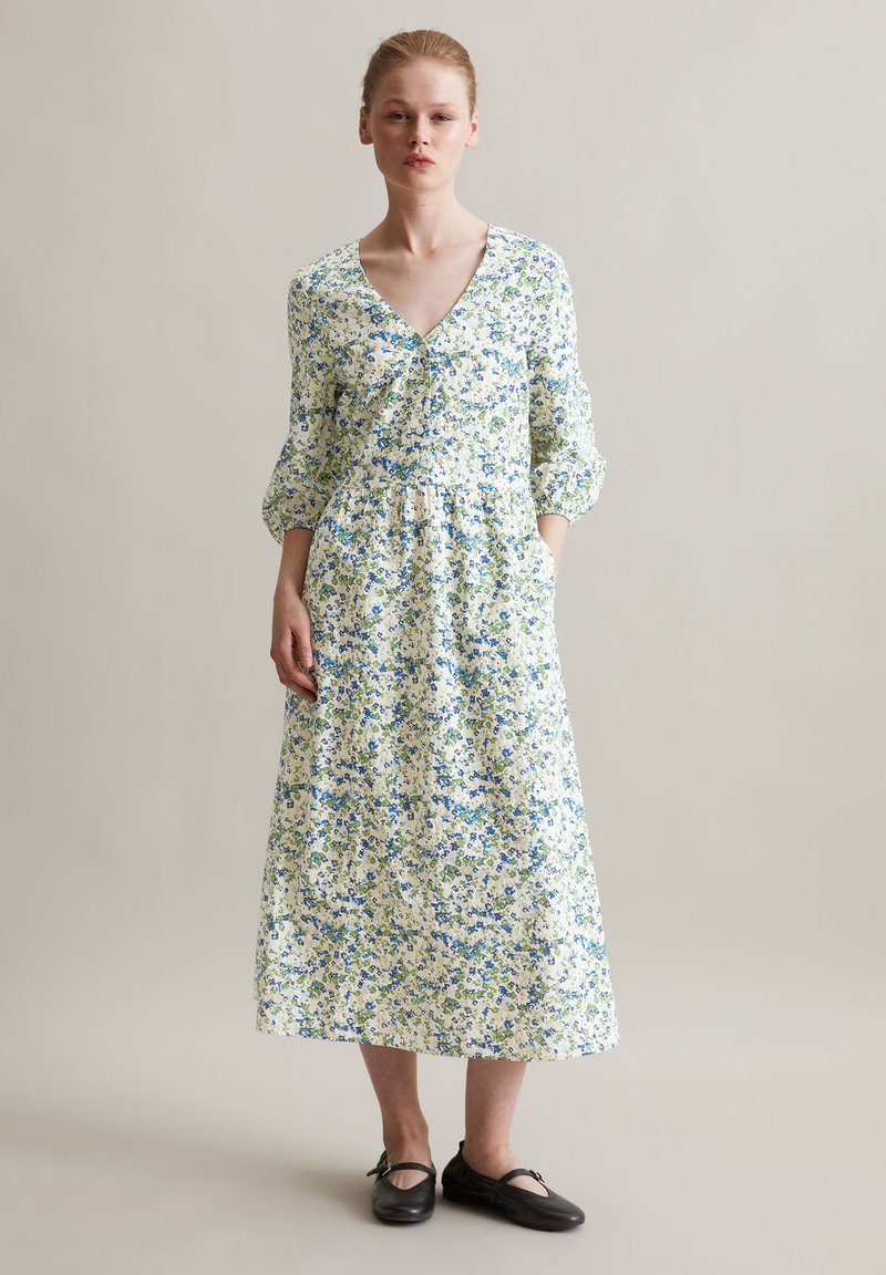 Das florale Midikleid hat einen V-Ausschnitt, dreiviertellange Ärmel und eine geraffte Taille. Der Stoff ist überwiegend weiß mit blauen und grünen Mustern.