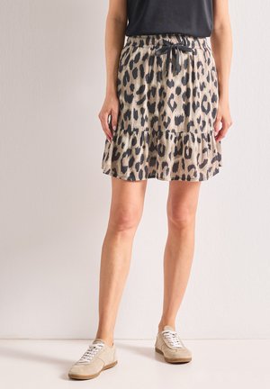 Femme portant une jupe beige et noire à imprimé léopard avec un ourlet à volants, un haut noir et des baskets beiges à lacets, debout contre un mur blanc.