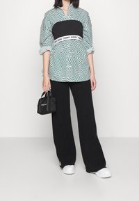 Zielona i biała koszula oversize w wzory, z czarnym pasem, w połączeniu z szerokimi czarnymi spodniami i małą czarną torbą, która ma białe akcenty z brandingiem.