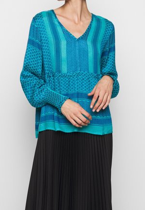 Kobieta ubrana w turkusowy, wzorzysty top z długim rękawem i dekoltem w serek oraz czarną plisowaną spódnicę, ręce złączone na wysokości talii.