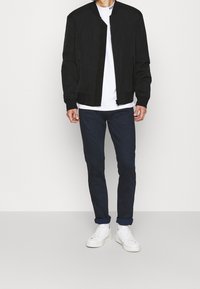 Giacca bomber nera con chiusura a zip, abbinata a una t-shirt bianca e pantaloni blu navy. L'outfit è completato da sneakers bianche.