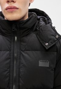 Crna jakna s podstavom i visokim ovratnikom, zatvaranje na zip, te epaulete na ramenima. Sadrži oznaku "HUGO" na prsima. Teksturedna tkanina.