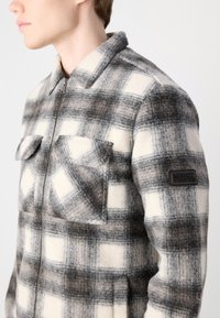Checkered wool jacket in gray, white, and black. Features a zip front, two chest pockets, and a branded patch on the left sleeve.