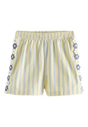 Enfant portant un short rayé jaune avec bordure florale, un haut à carreaux volanté et des sandales blanches à boucle, debout sur un fond clair.