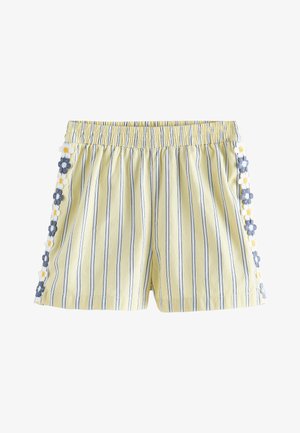 Kind draagt gele gestreepte shorts met bloemige rand, een gerimpelde geruite top en witte sandalen met gesp, staande op een lichte achtergrond.