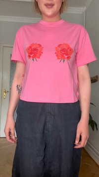 T-shirt cropped de algodão rosa com dois gráficos de rosas vermelhas na frente. As mangas são curtas, com um ajuste relaxado e textura suave.