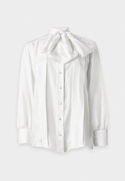 Blouse blanche à manches longues avec boutons sur le devant et un grand nœud noué au col, avec des détails plissés à l'avant.