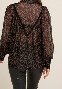 Blouse noire transparente avec motif abstrait brun foncé, détail en dentelle en forme de V dans le dos, manches longues et poignets à boutons.