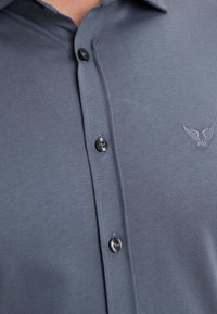 Primer plano de camisa azul grisácea con botones y pequeño logo bordado con forma de ala en el pecho cerca de los botones.