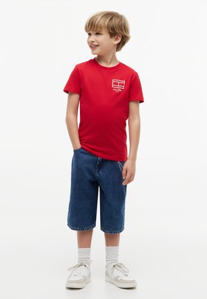 Garçon souriant, portant un t-shirt rouge à manches courtes avec logo, un short en jean longueur genou, des chaussettes blanches et des baskets blanches, debout avec une main dans la poche.