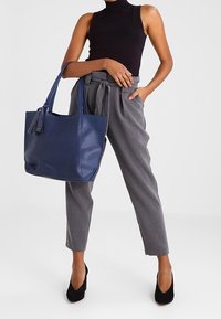 Námořnická modrá taška přes rameno s perforovaným designem, hladkou texturou a dlouhými uchy, kombinovaná se šedými nadměrnými kalhotami a černými podpatky.