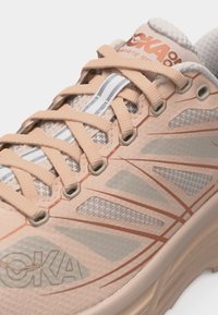 Prim-plan cu pantof de alergare bej și gri deschis, cu șireturi încrucișate, plasă respirabilă și talpă groasă și amortizată.